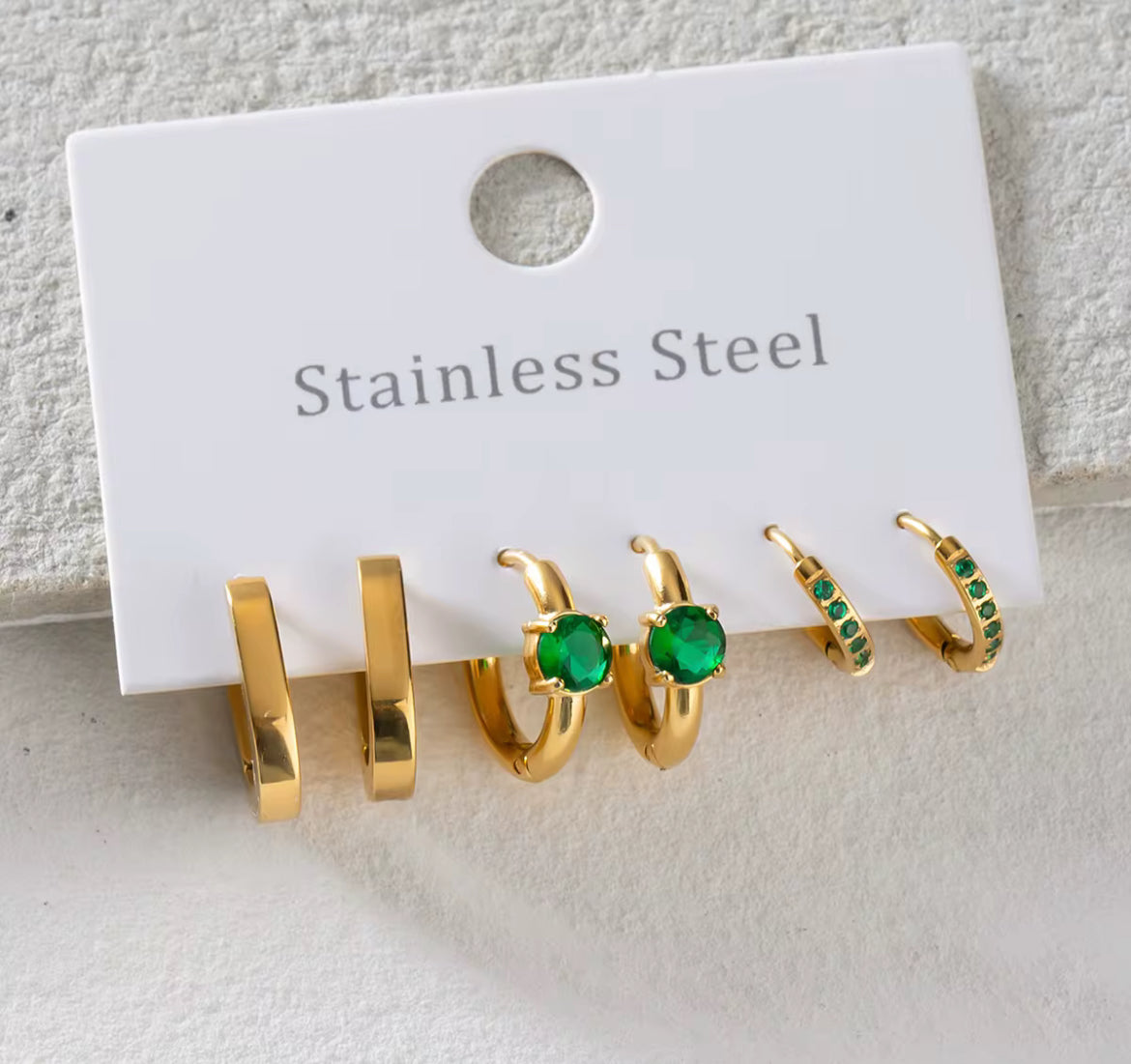 Set of gold hoop earrings with green gemstones on a white card labeled 'Stainless Steel'.