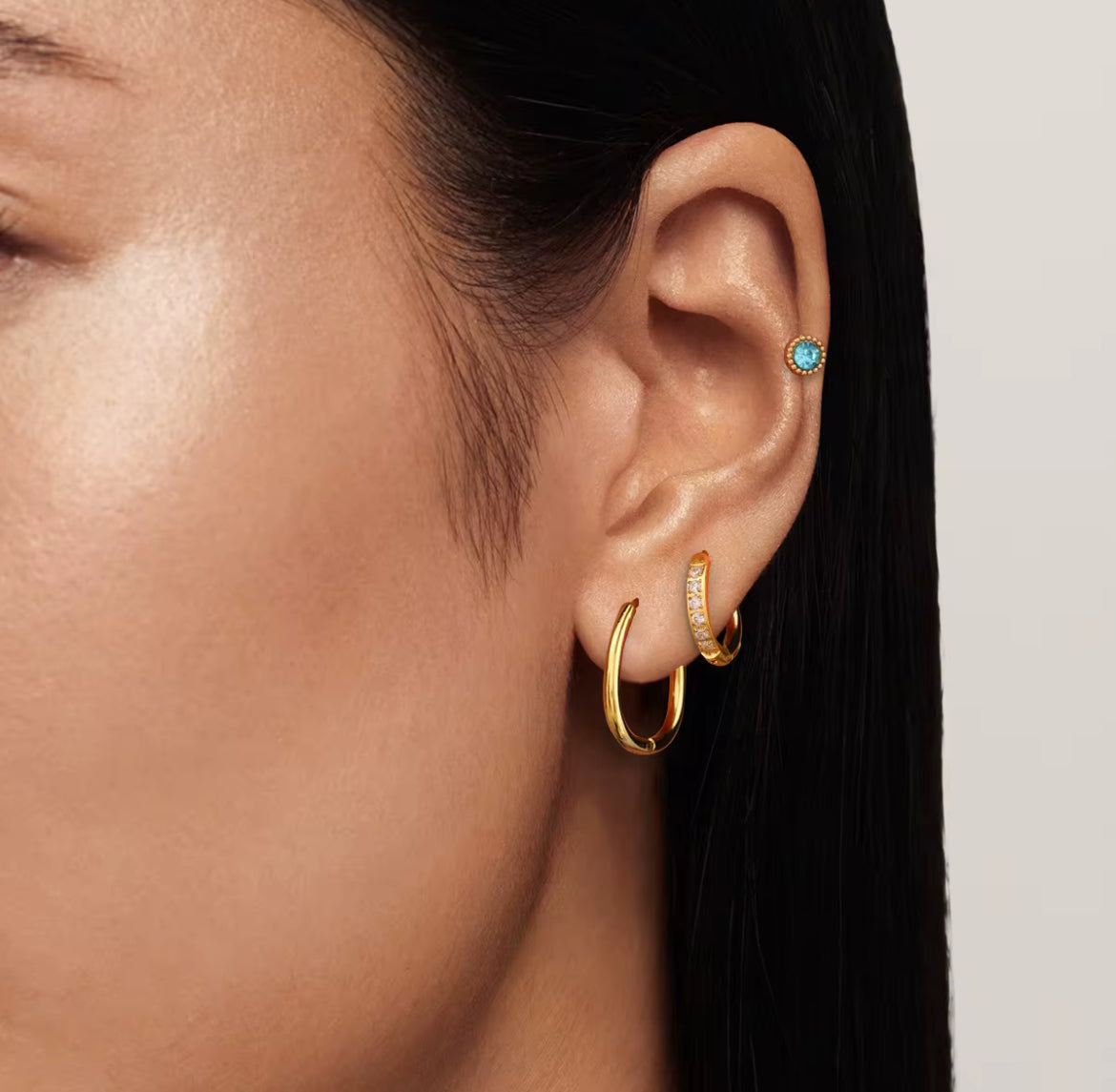 Close-up of an ear wearing gold hoop earrings with gemstones on a neutral background