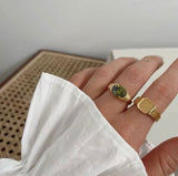 Hand wearing two gold rings with a green gemstone on a neutral background