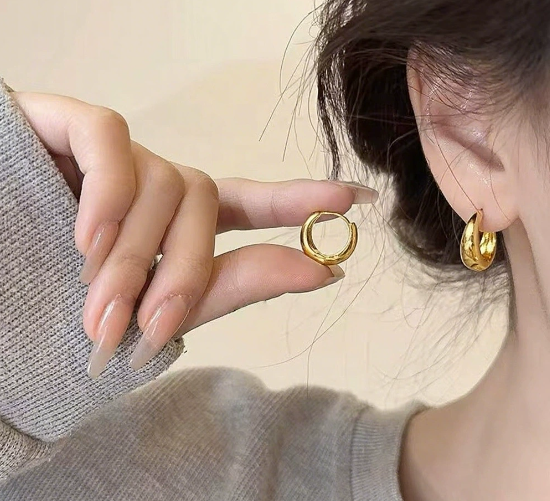 Gold hoop earrings being worn and held by a hand against a neutral background
