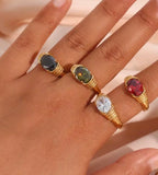 Hand wearing four gold rings with different colored gemstones on a neutral background